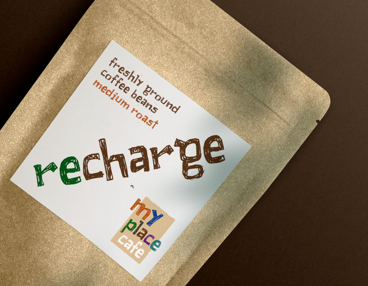 A paper bag of freshly ground coffee beans with 'Recharge' branding in logo font and the cafe logo